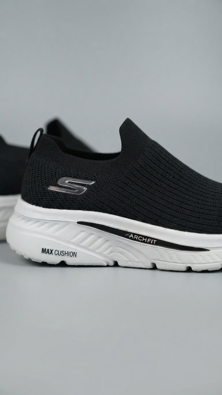 A black slip-on knit sneaker by Skechers with a white sole, featuring the Skechers logo on the side and 'ARCHFIT' and 'MAX CUSHION' text on the midsole, shown against a neutral gray background.