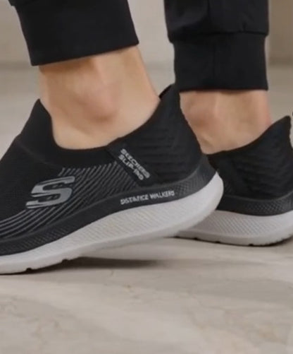 Close-up of a person wearing black Skechers Distance Walkers slip-on sneakers with a white sole, standing on a light-colored tiled floor.
